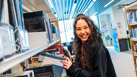 A student at The Manchester College is taking a library book off a shelf. She is looking past the camera and smiling.