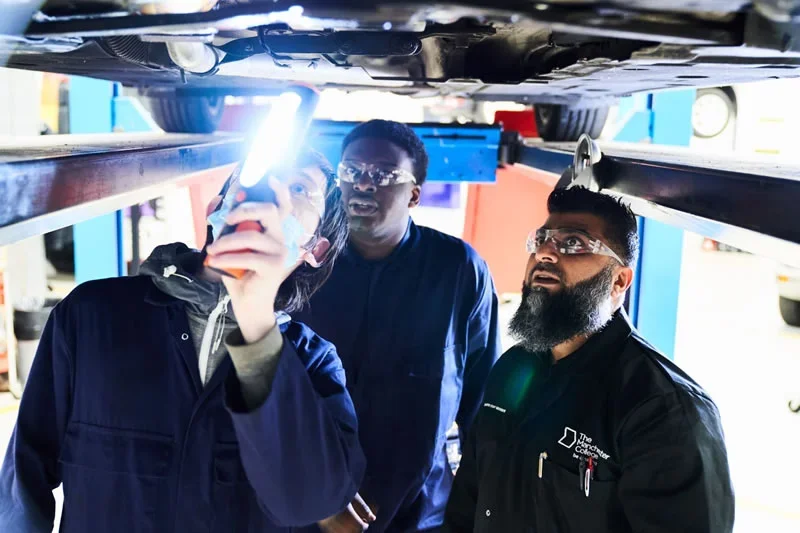 College learners in workshop overalls and safety glasses inspecting the underside of a raised vehicle, with one using a handheld light.
